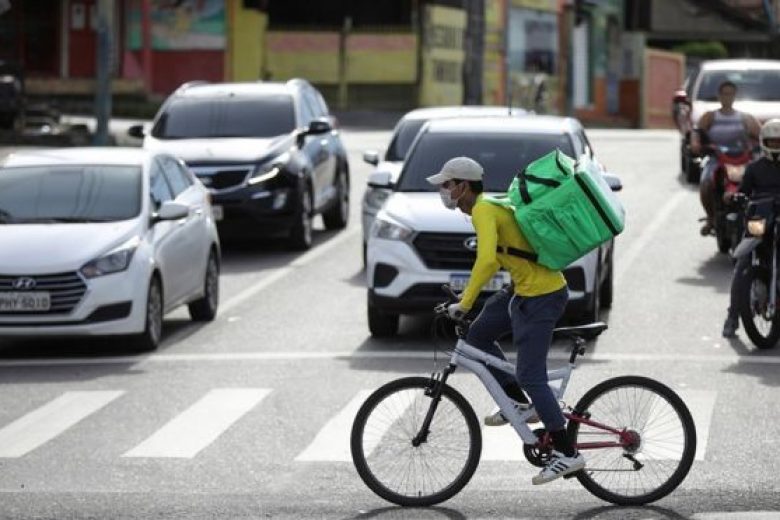 Foto de entregador de aplicativo andando de bicicleta em uma faixa zebrada em frente a carros